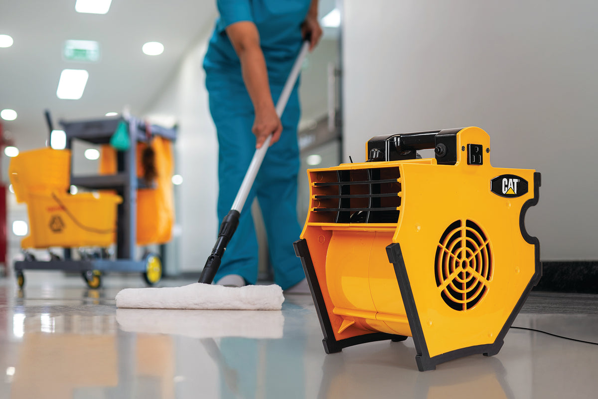 A yellow CAT floor drying machine in the foreground, with a person in blue cleaning uniforms mopping a hallway floor in the background.