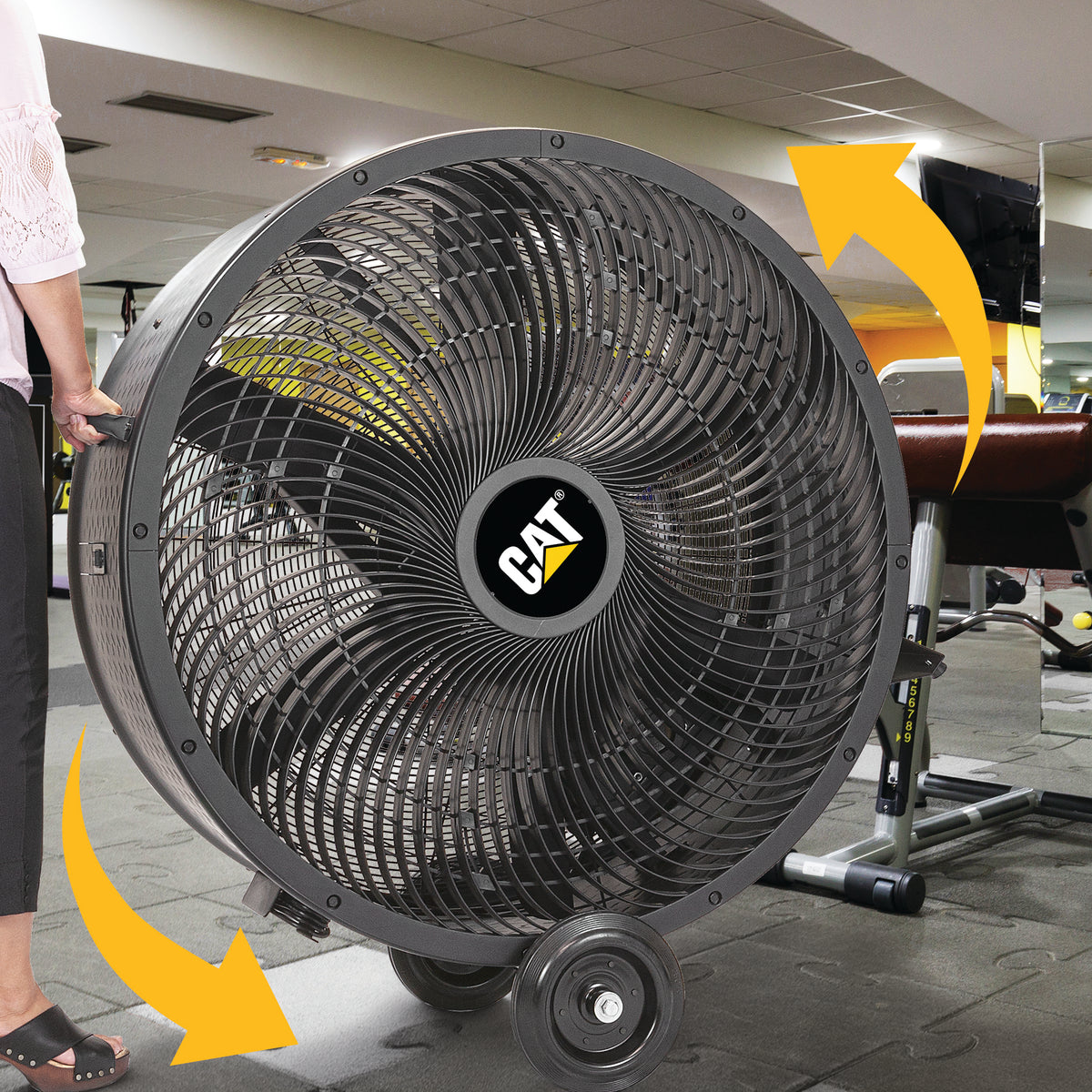 A large CAT industrial floor fan with wheels, shown in a gym setting with a person's hand and foot visible on the left and yellow arrows indicating airflow direction.