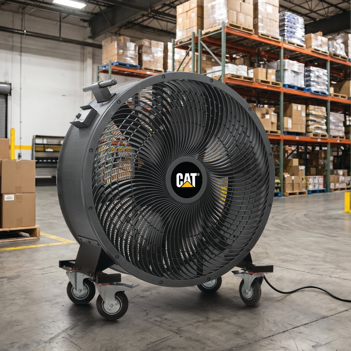 A large CAT industrial floor fan with a black protective grille and caster wheels, positioned in a warehouse setting with shelving and boxes in the background.