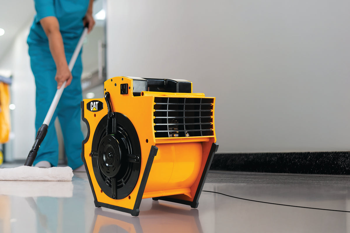 A yellow and black CAT industrial floor fan placed on a glossy floor in a clean indoor setting, with a person in teal cleaning attire mopping in the background.