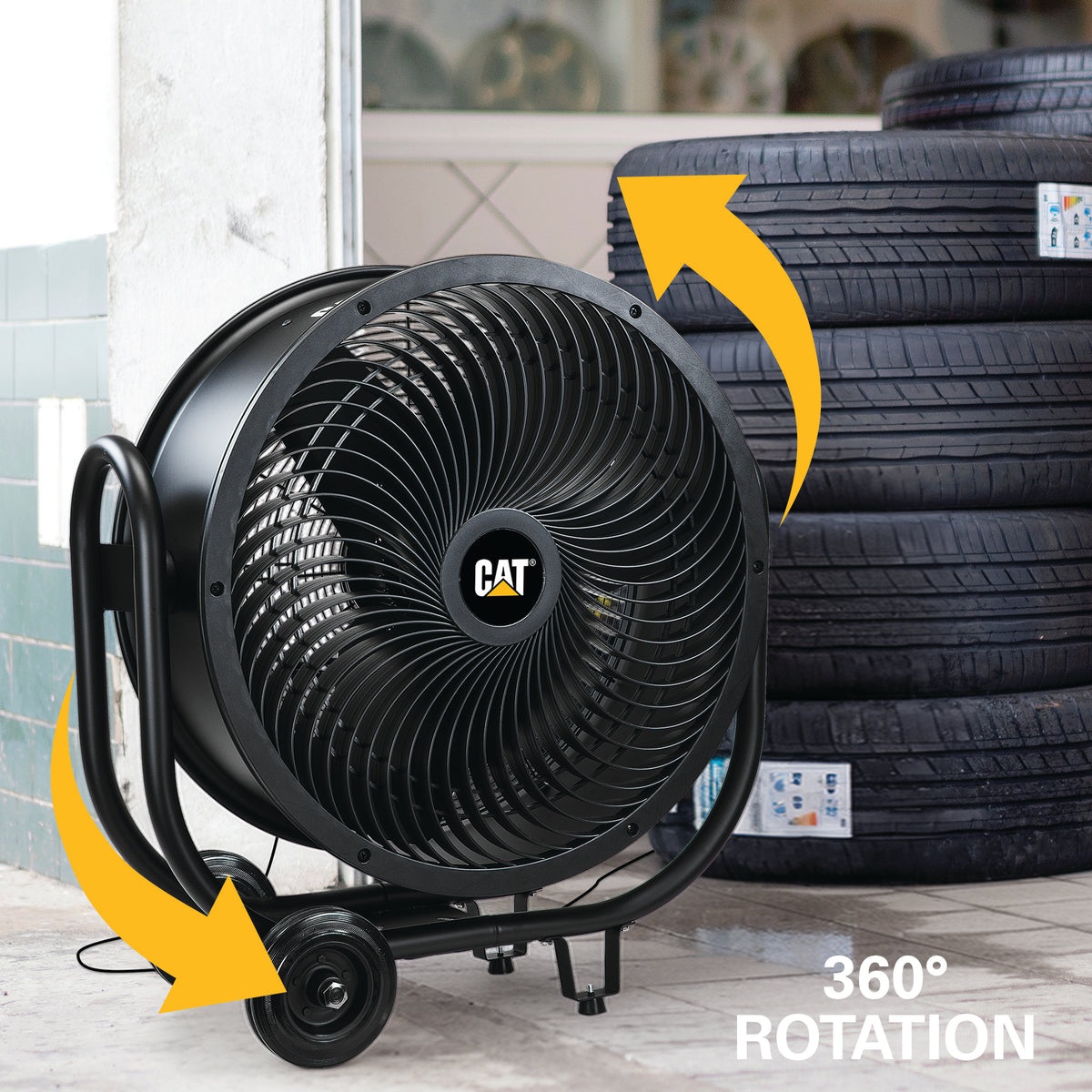 A black CAT industrial floor fan with a 360-degree rotation feature, shown in a garage-like setting with yellow arrows indicating movement and tires in the background.