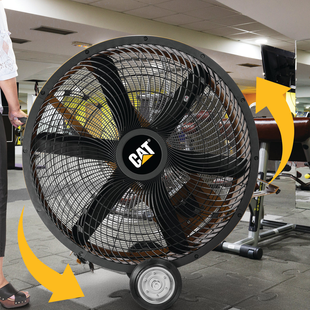 A large CAT industrial floor fan with a black metal grille and black blades, shown in a gym setting with yellow motion arrows indicating airflow direction. A person's leg is visible on the left.