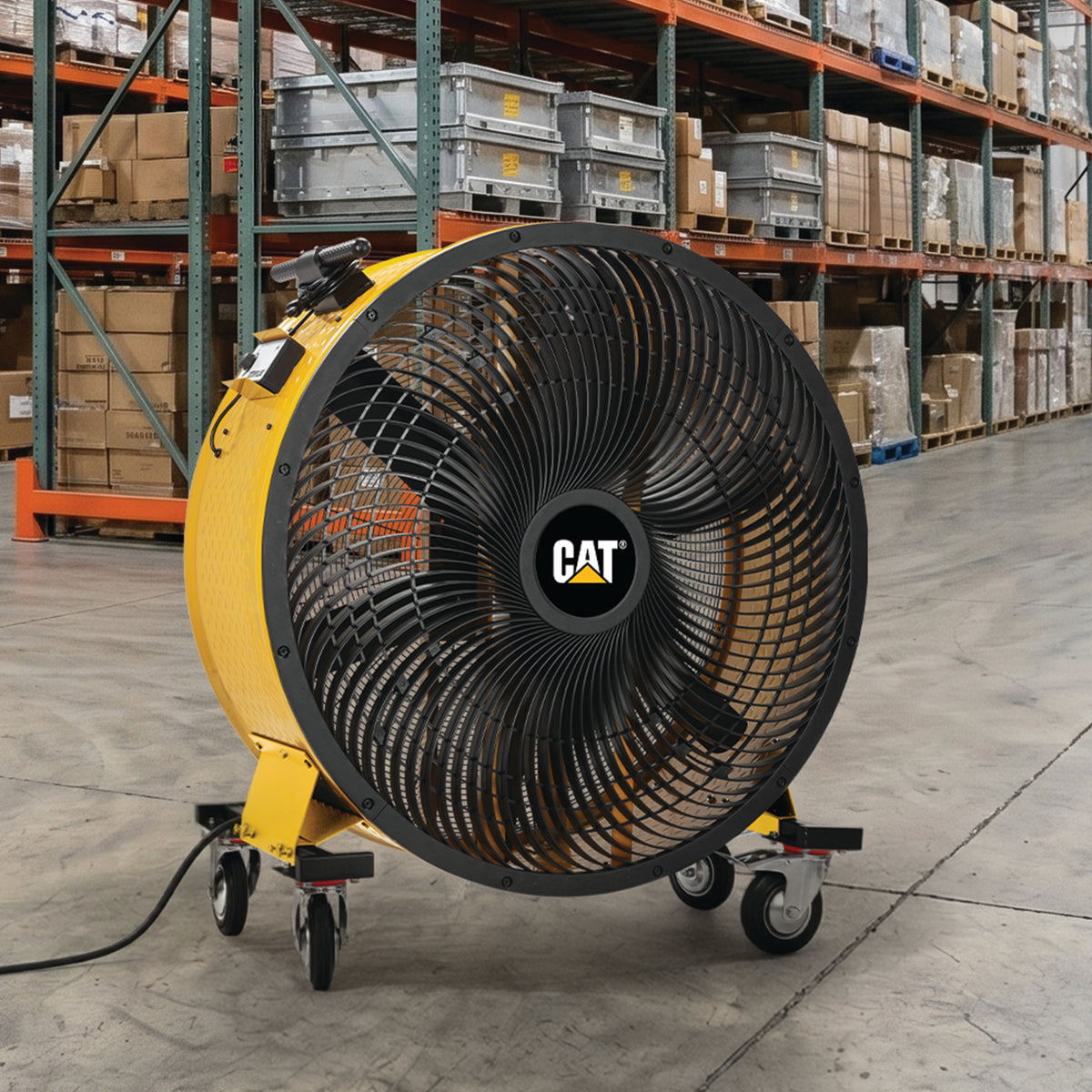 A large yellow and black CAT industrial fan with a protective grille and caster wheels, positioned in a warehouse setting with shelving and boxes in the background.
